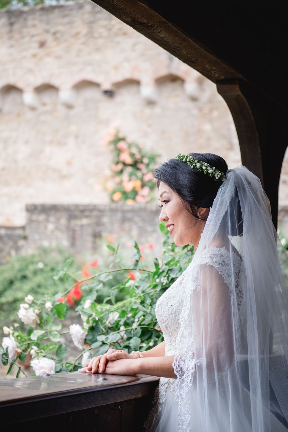 A bride in a lace veil stands at a rustic window overlooking a garden with blooming flowers.