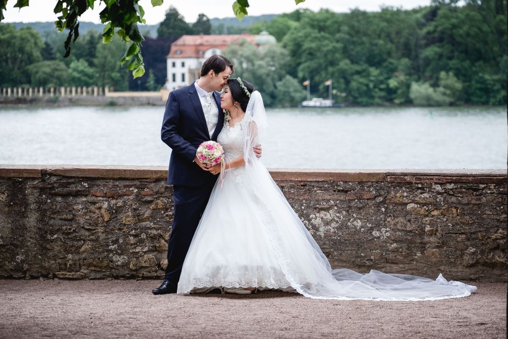 A couple in wedding attire stands by a stone wall overlooking a serene lake.