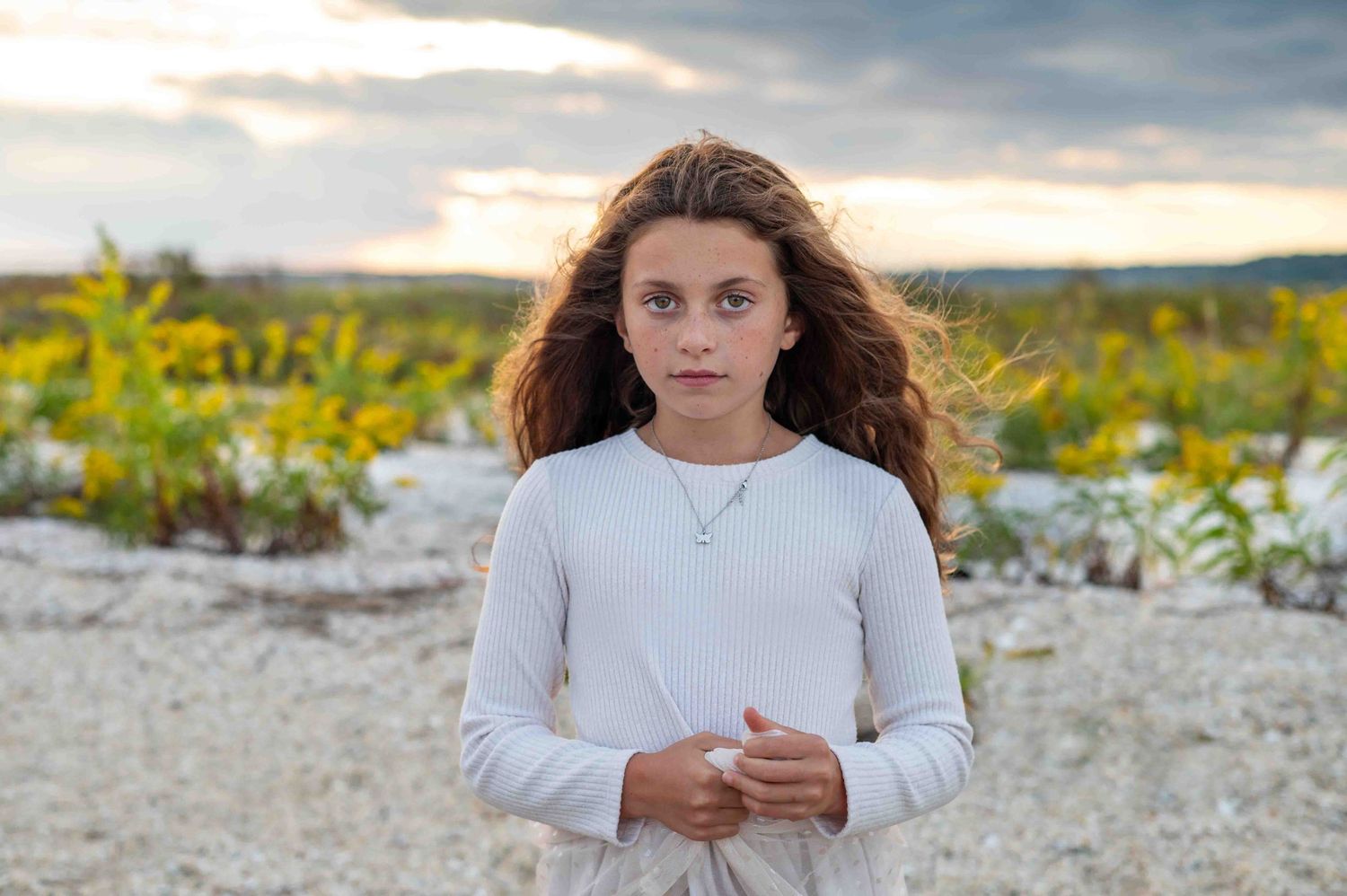 A young girl in a white sweater stands on rocky terrain with yellow flowers and a cloudy sky in the background.