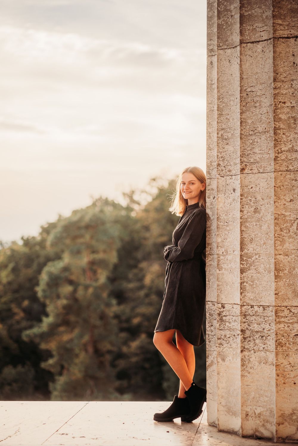 Woman in a black dress poses elegantly against a stone column with nature backdrop.
