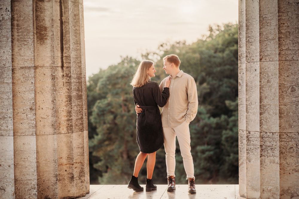 Couple standing between stone columns at sunset in elegant outfits.