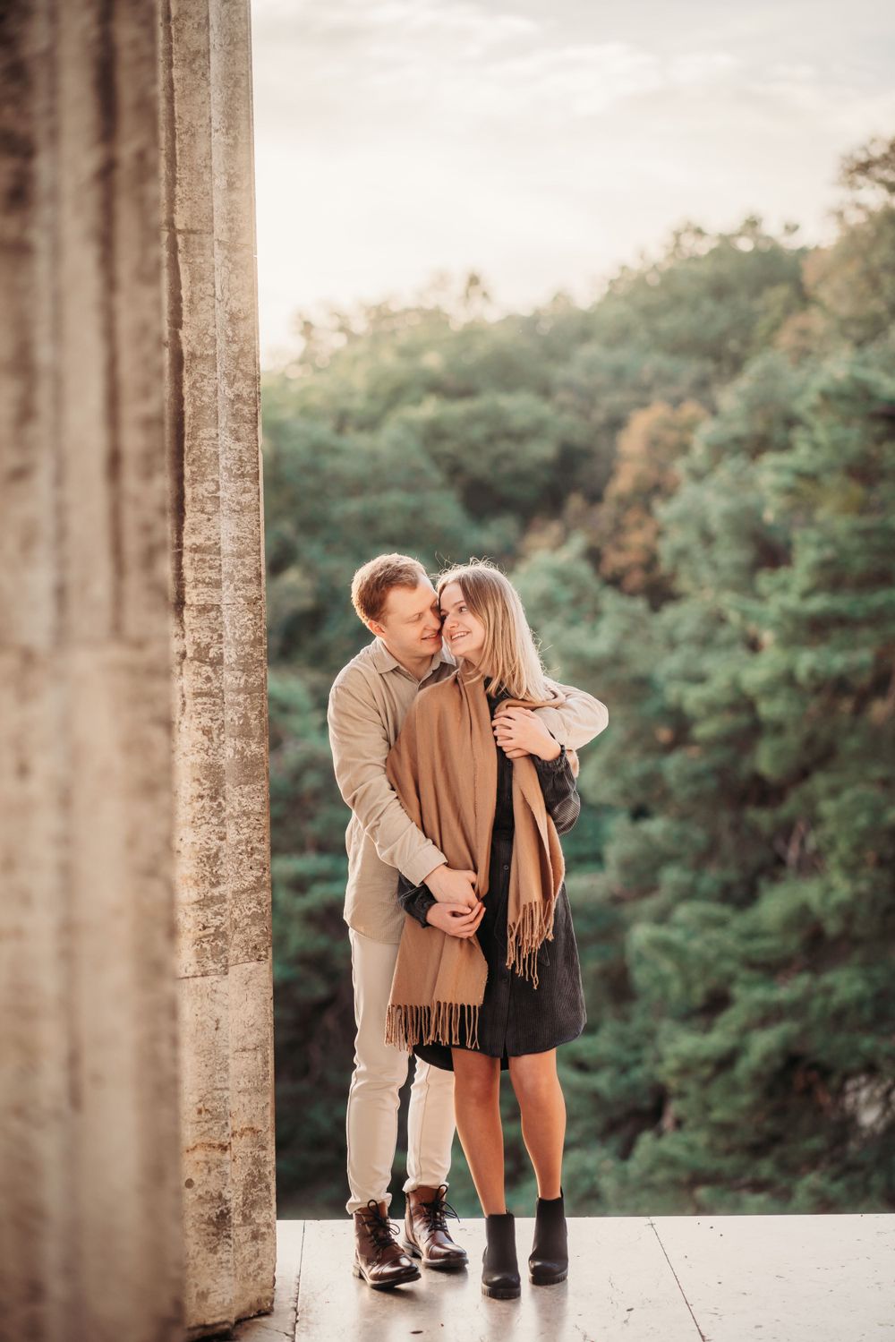 A romantic couple shares an intimate moment against stone pillars with a scenic forest backdrop.