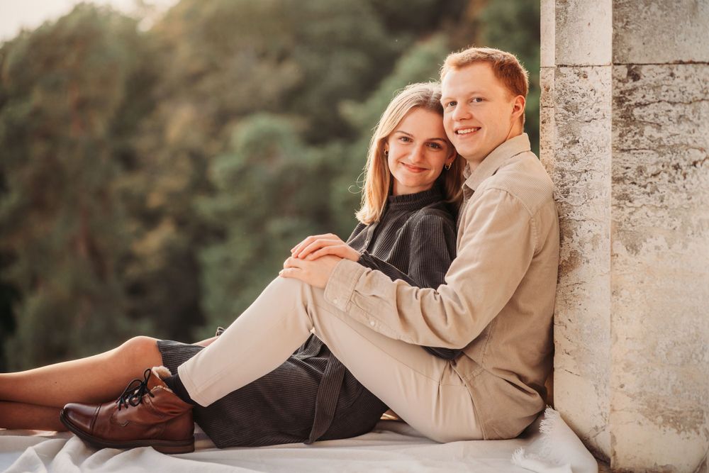 A couple in casual attire sits together on stone ledge with greenery in background.