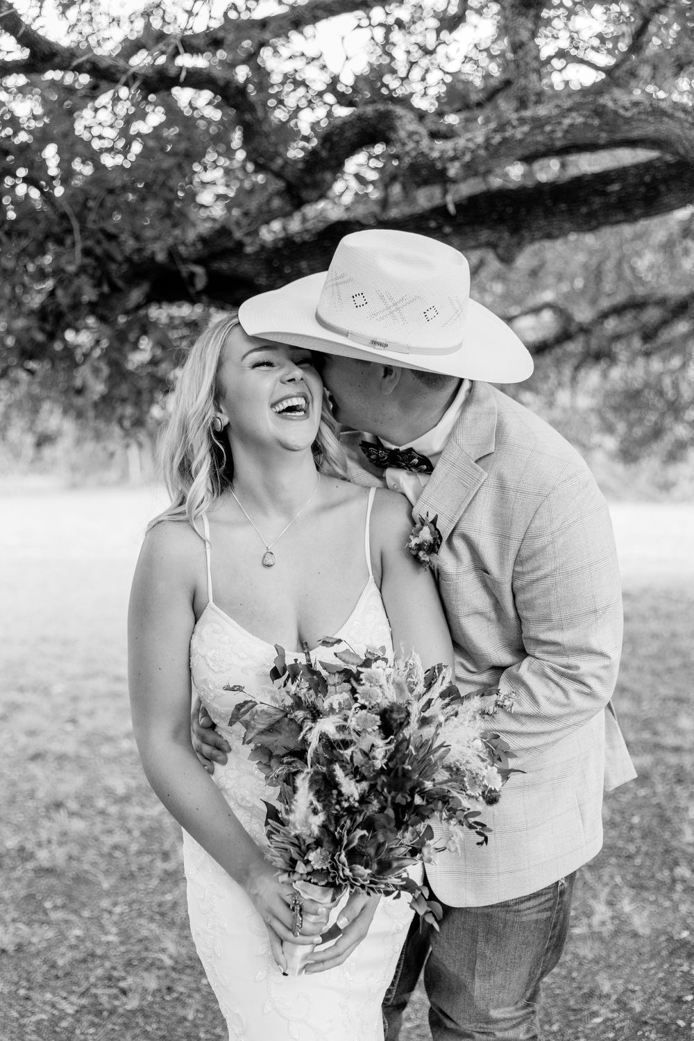 Joyful wedding couple laughing together under tree branches wearing country western attire.