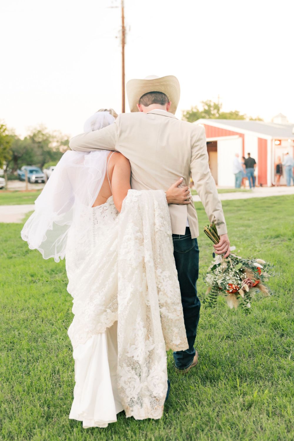 Elegant bride in lace dress and cowboy-styled groom dance together on grassy field.