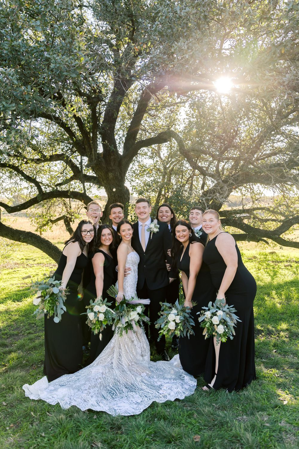 Bridal party in black dresses and white wedding gown sit together under a sunlit tree during outdoor wedding photos.