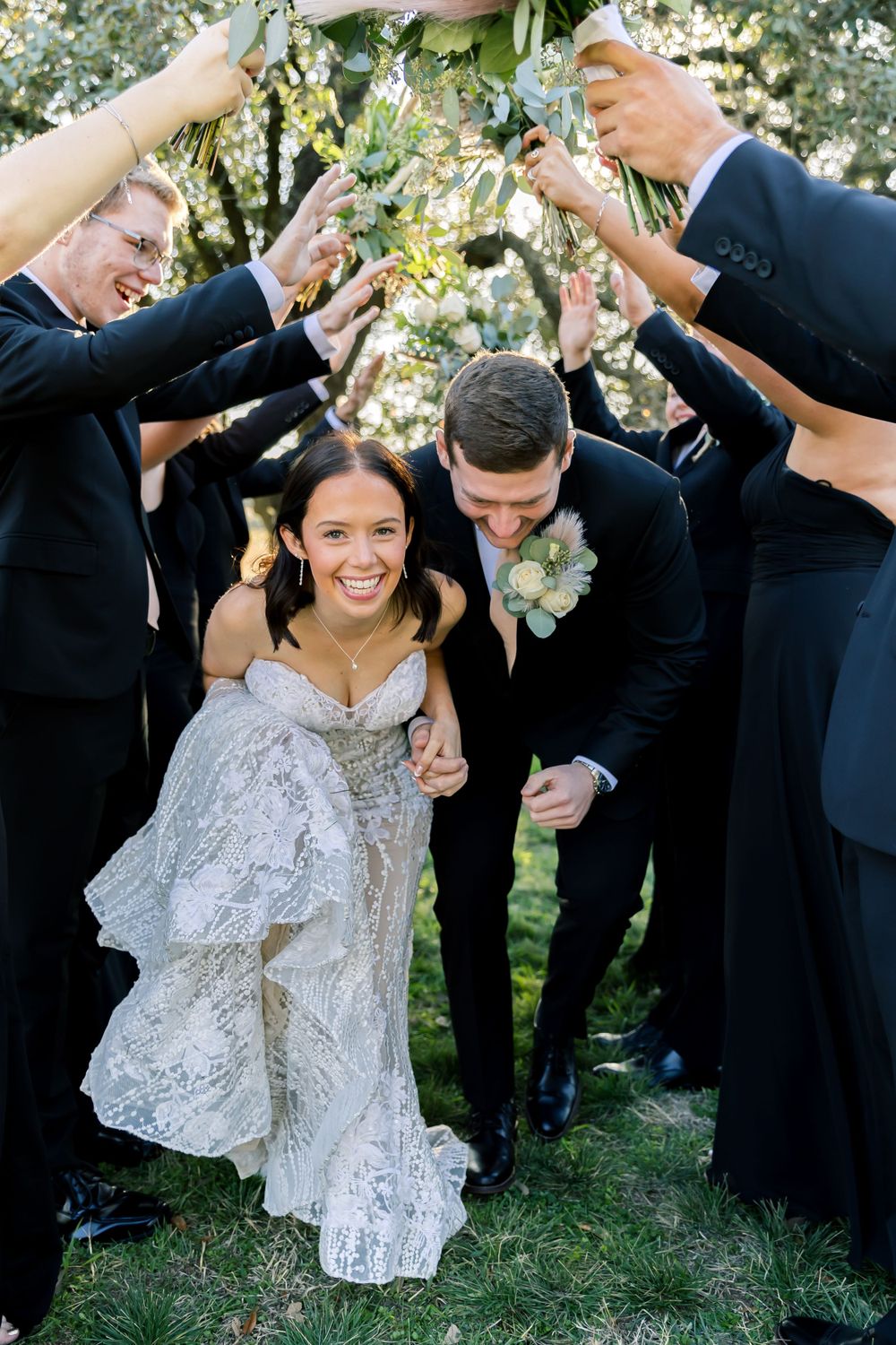 Wedding party group of groomsmen in black suits huddles around a bride in white lace dress in outdoor garden setting.