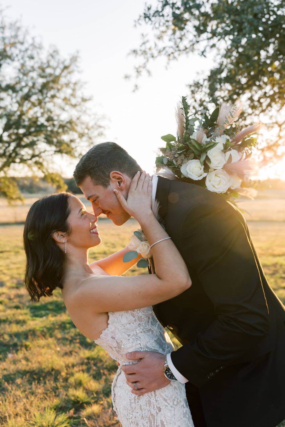 A romantic wedding couple embraces in a golden sunlit field with a bouquet of white flowers during sunset.
