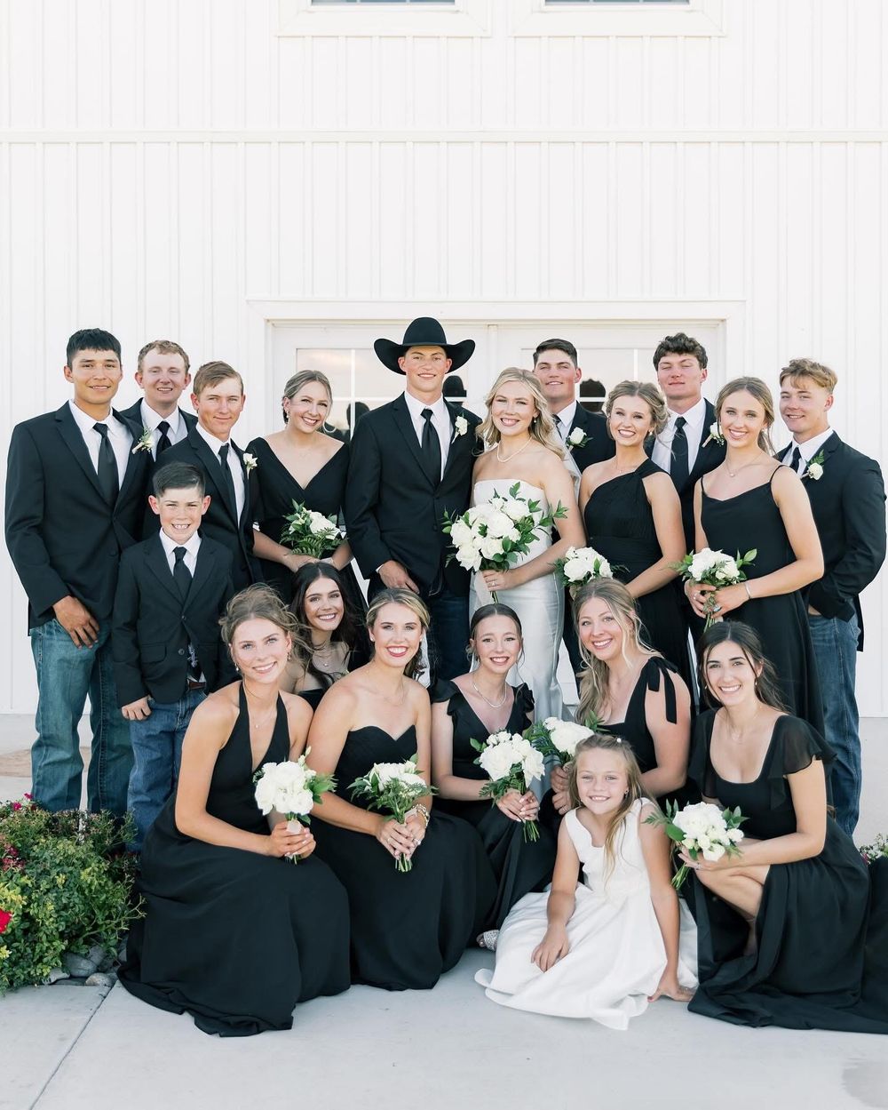 Wedding party in black formal attire poses together against white building backdrop.
