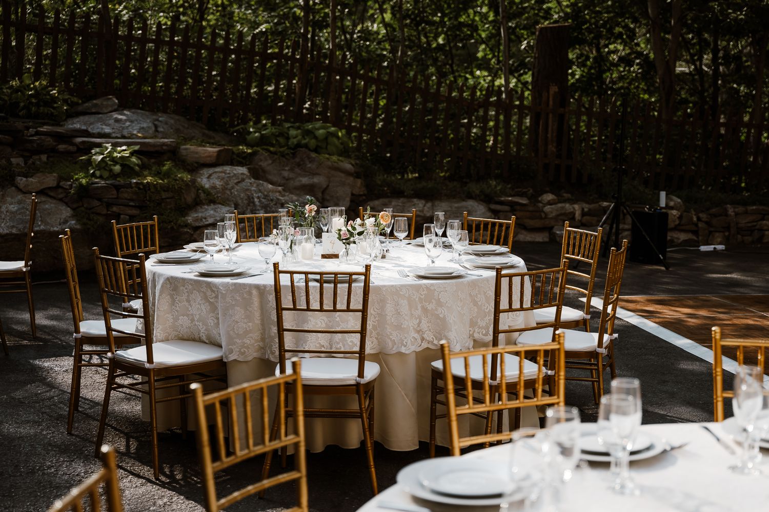 Elegant outdoor wedding reception table setting with white linens and wooden chiavari chairs in a garden venue.
