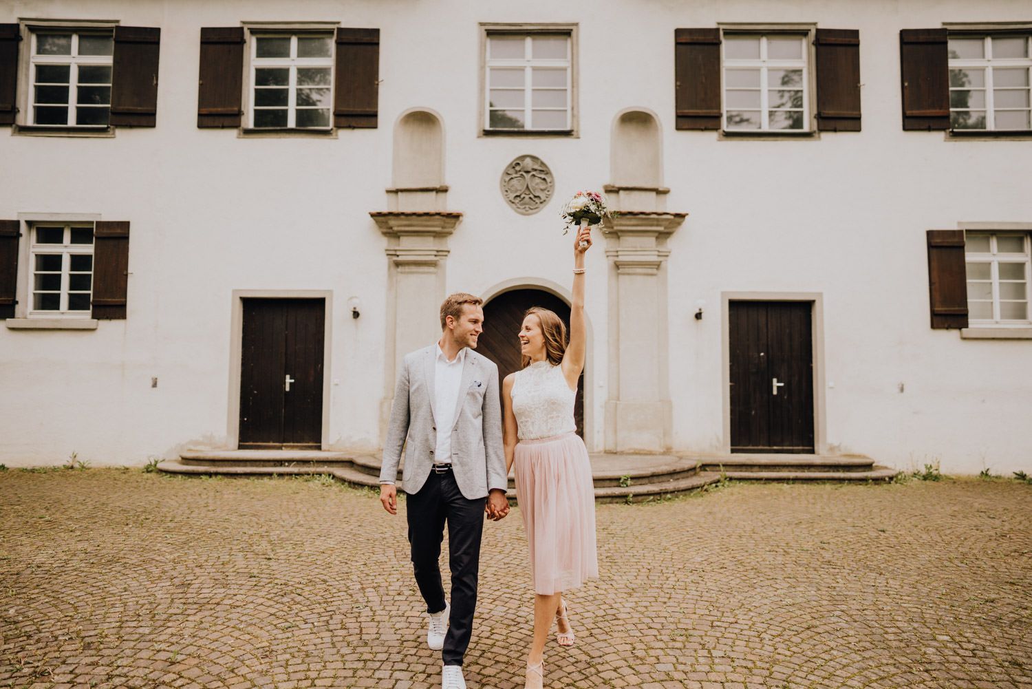A couple stands together on cobblestones before a classic white building with symmetrical windows.