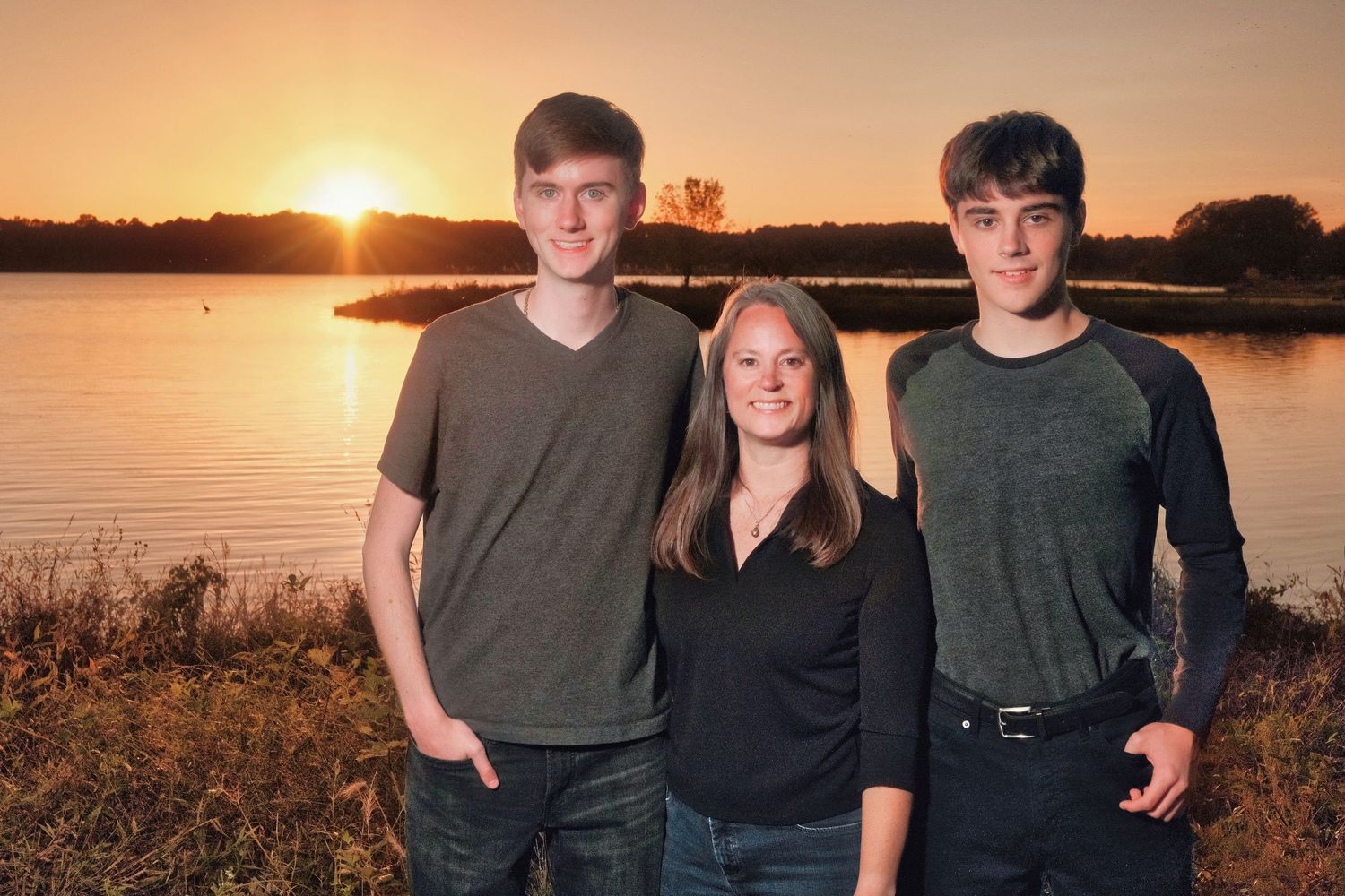 Three people posing together at sunset by a lake with golden sunlight reflecting on the water.