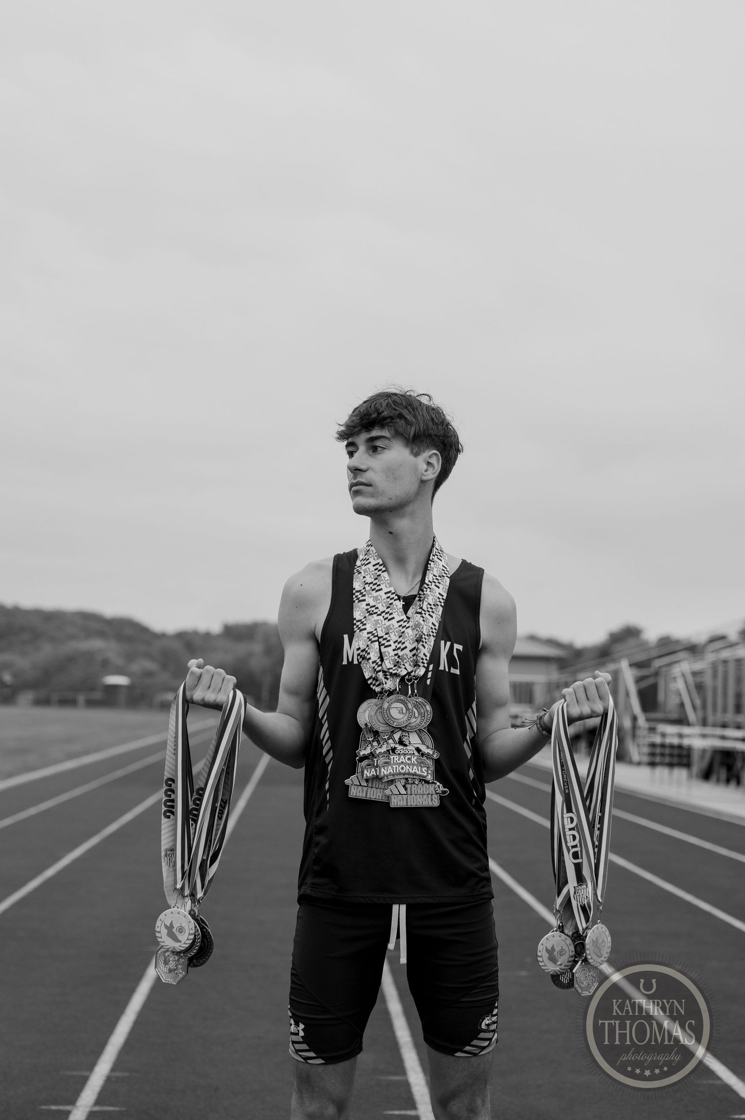 Black and white portrait of athlete holding medals on track field with overcast sky.