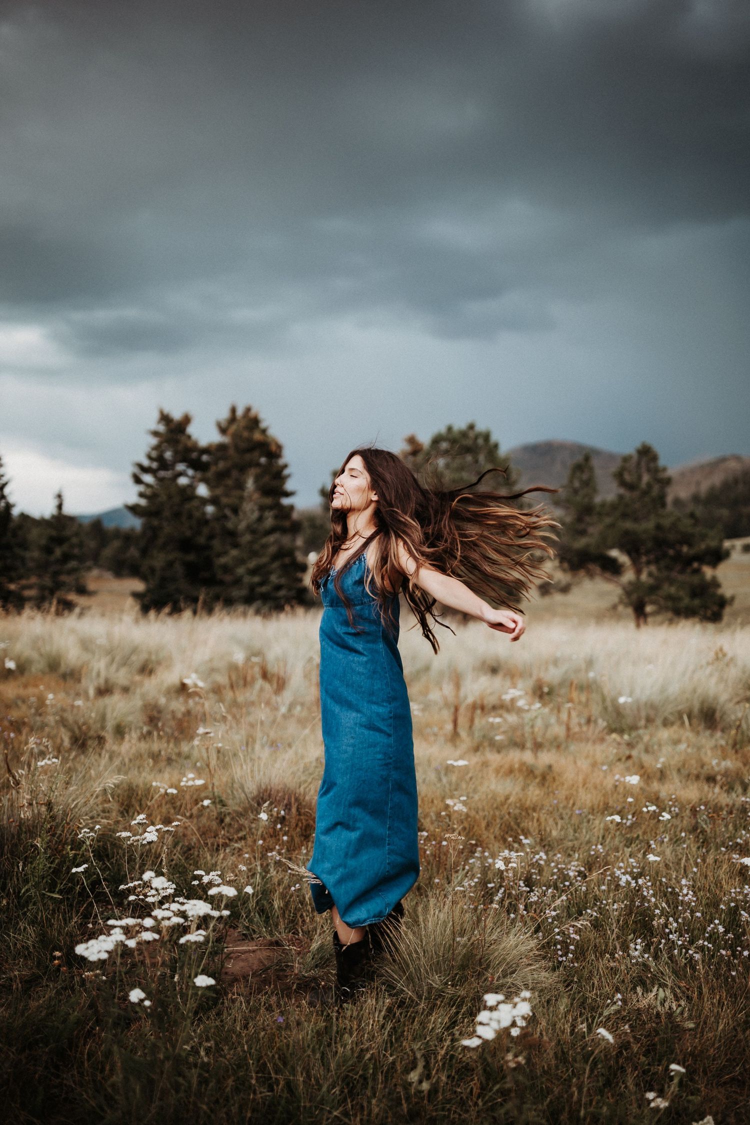 Woman with flowing hair in blue dress stands among wildflowers in mountain field.
