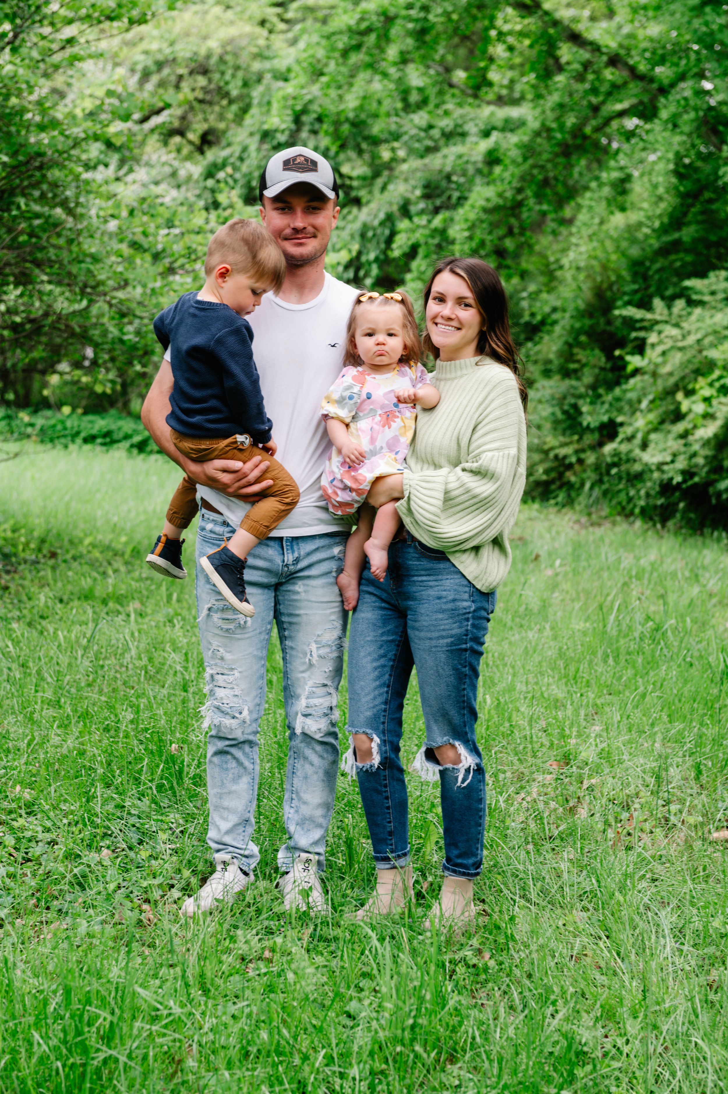 A family stands together on green grass with trees in the background wearing casual clothing on a spring day.