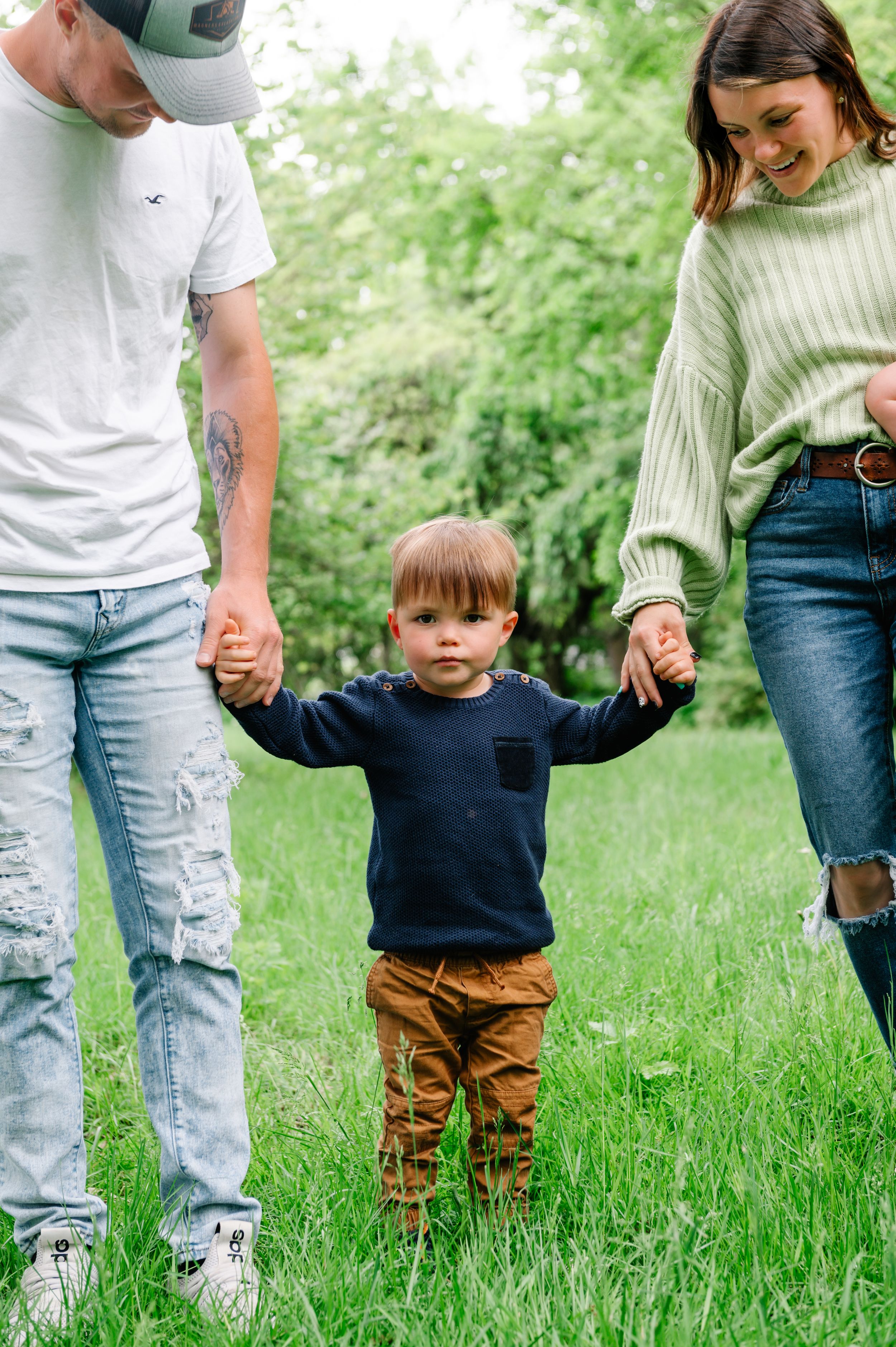 A toddler in a navy sweater and brown pants holds hands with adults while standing in green grass on a sunny day.