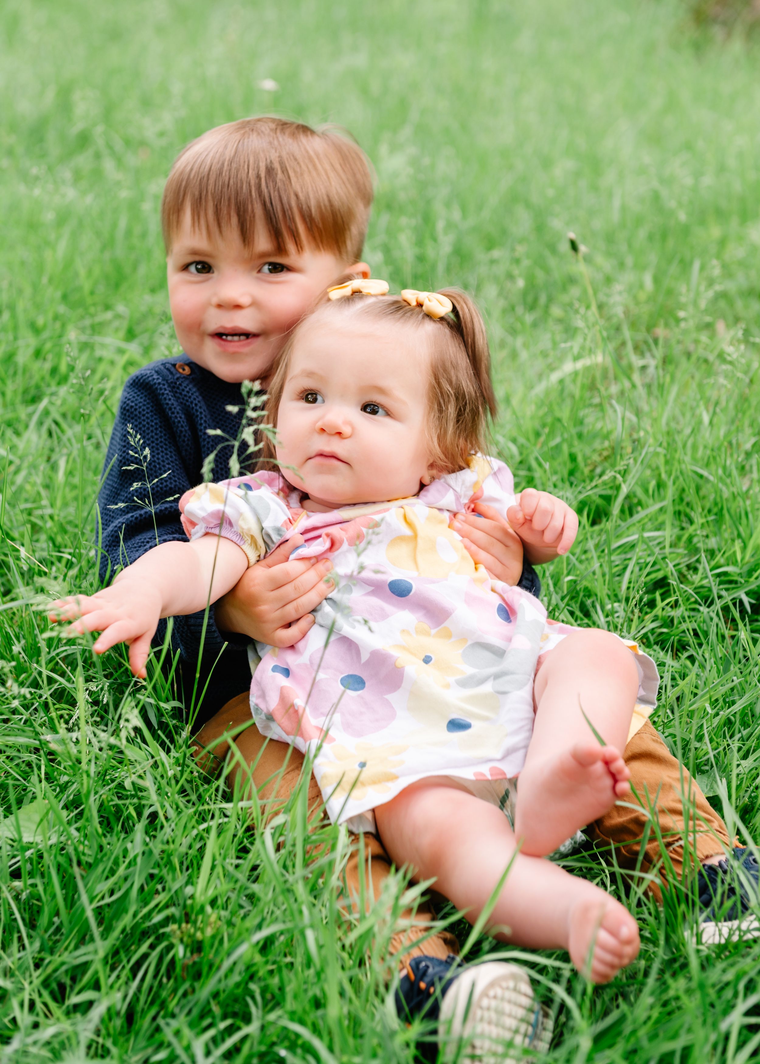 Two children sit together in green grass on a sunny day, with the older one holding the younger one.