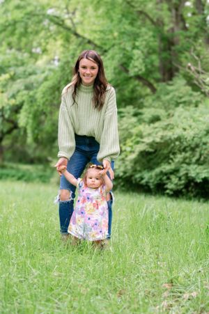 A young toddler in a floral dress stands in a grassy meadow with adult guidance on a spring day.