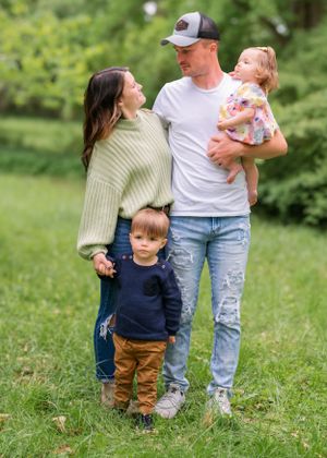 A family stands together on green grass, wearing casual clothing and smiling outdoors in a natural setting.