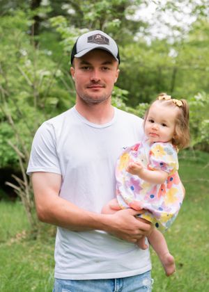 A person in a baseball cap holds a toddler wearing a colorful floral dress against a green outdoor backdrop.