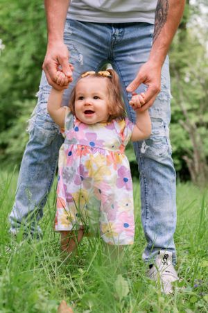 A small child in a colorful floral dress learns to walk while holding hands in a grassy outdoor setting.