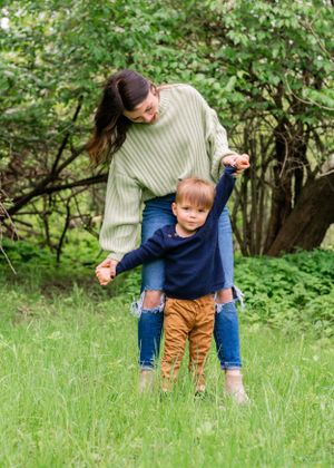 A parent helps a small child learn to walk through tall grass in a wooded outdoor setting on a sunny day.