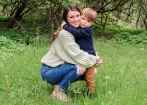 A person in a cream sweater and jeans crouches in green grass while hugging a small child wearing navy blue.
