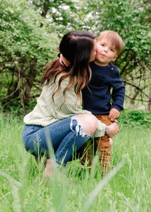 A tender outdoor moment between parent and toddler sitting in green grass surrounded by leafy foliage.