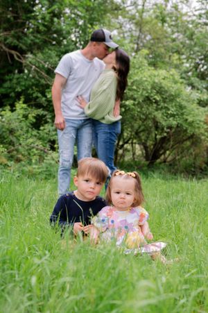 Family portrait in green meadow with toddlers sitting in tall grass while couple embraces behind them.