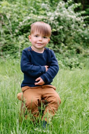 Small child wearing navy sweater and brown pants sits in tall grass surrounded by flowering bushes in a garden setting.