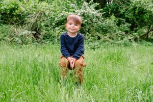 Young child in a navy sweater and brown pants sits happily in tall grass with green foliage in the background.