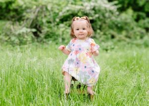 Young child wearing a floral dress stands in a field of tall grass with blooming wildflowers in the background.