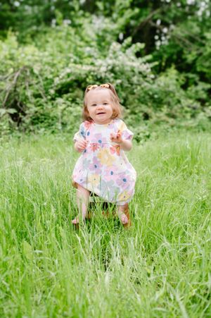 A toddler wearing a floral dress smiles joyfully while standing in tall grass surrounded by green trees.