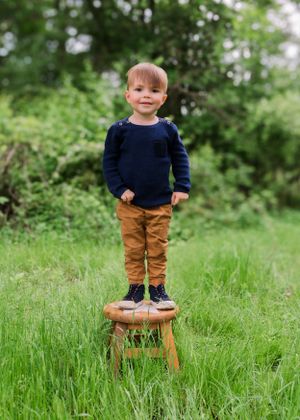 Small child in navy sweater and brown pants stands confidently on wooden stool in lush green outdoor setting.