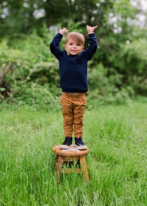 Small child in navy sweater and brown pants stands on wooden stool in lush green grass with arms raised victoriously.