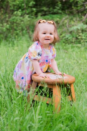 Small child in floral dress smiles while leaning on wooden stool amid tall green grass outdoors.