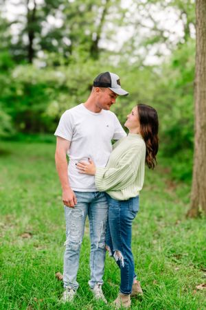 A couple stands embracing in a lush green park setting wearing casual outfits of denim and white.