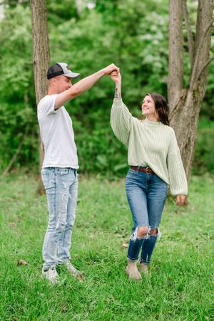 Two people in casual wear dancing and twirling together on green grass with trees in the background.
