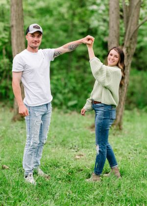A couple holding hands and posing together in a grassy outdoor setting with trees behind them.