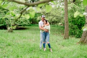 A couple embraces while standing on grass in a wooded area on a sunny day.