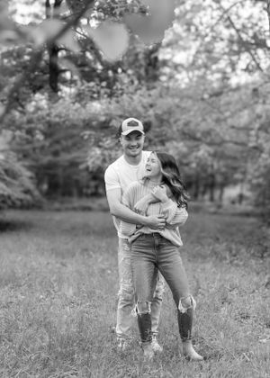 A black and white portrait of a couple embracing in a wooded meadow setting.