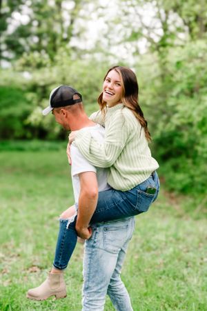 Playful couple sharing a piggyback ride outdoors in a green spring setting wearing casual jeans and sweaters.
