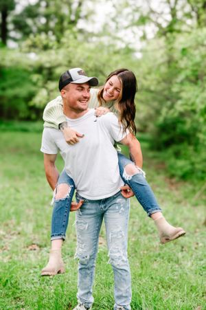 A playful couple enjoys a piggyback ride in a green spring meadow, wearing casual denim and t-shirts.