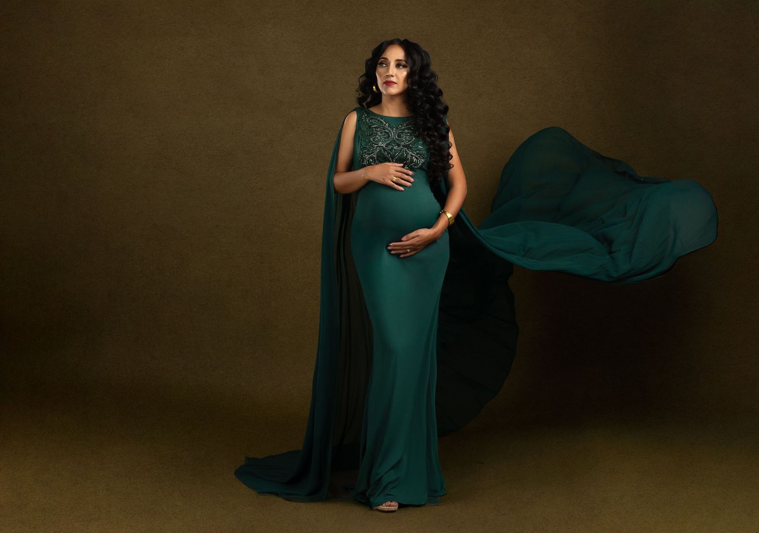 Elegant maternity photo featuring a flowing emerald green cape dress against a moody dark backdrop.