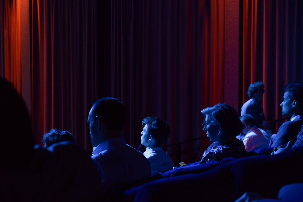 Audience members seated in dark theater seats illuminated by dramatic red lighting.