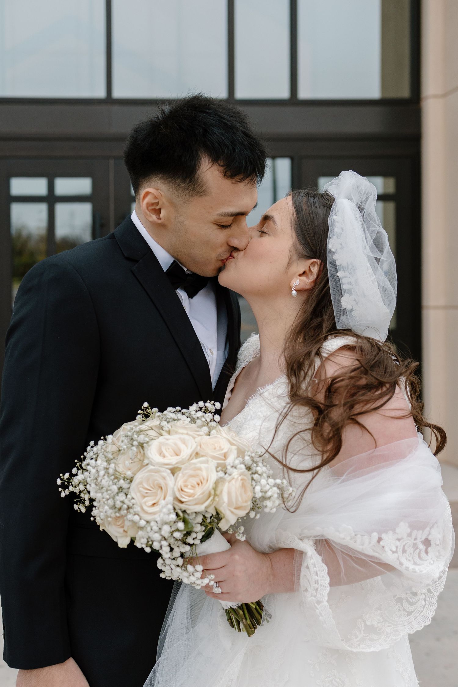 Newlyweds share a romantic kiss while holding a white rose and baby's breath bouquet.