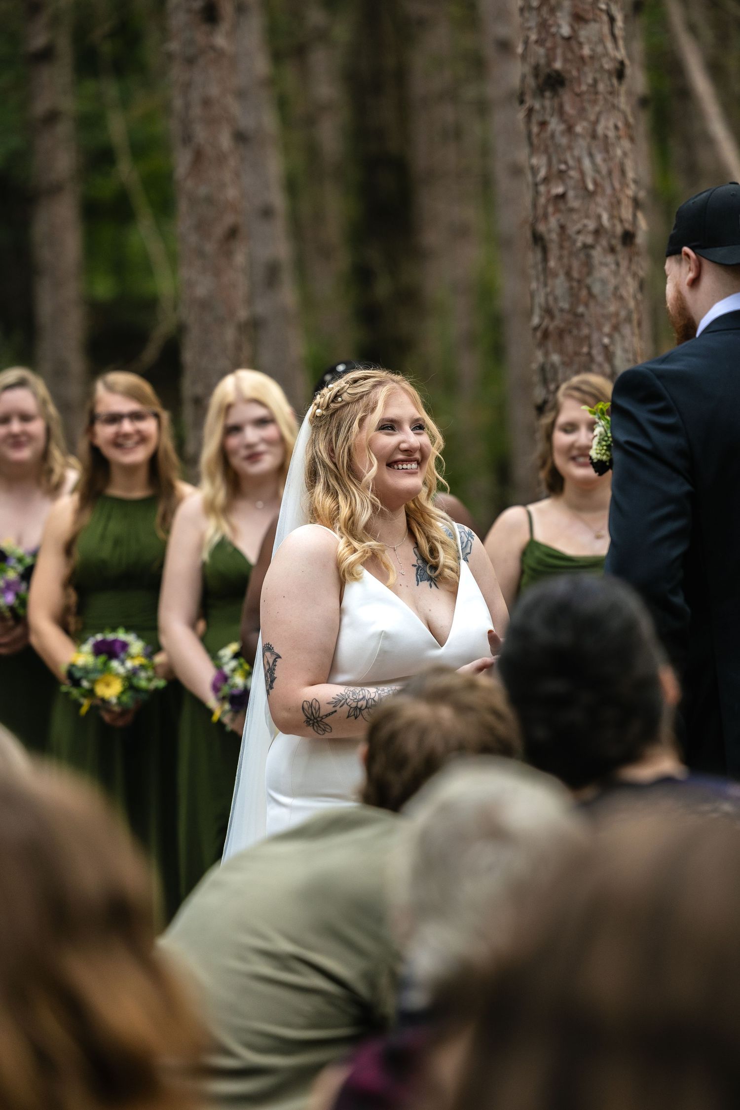 A joyful outdoor wedding ceremony in a forest with bridesmaids in dark green dresses holding colorful bouquets. Ludington, Michigan.