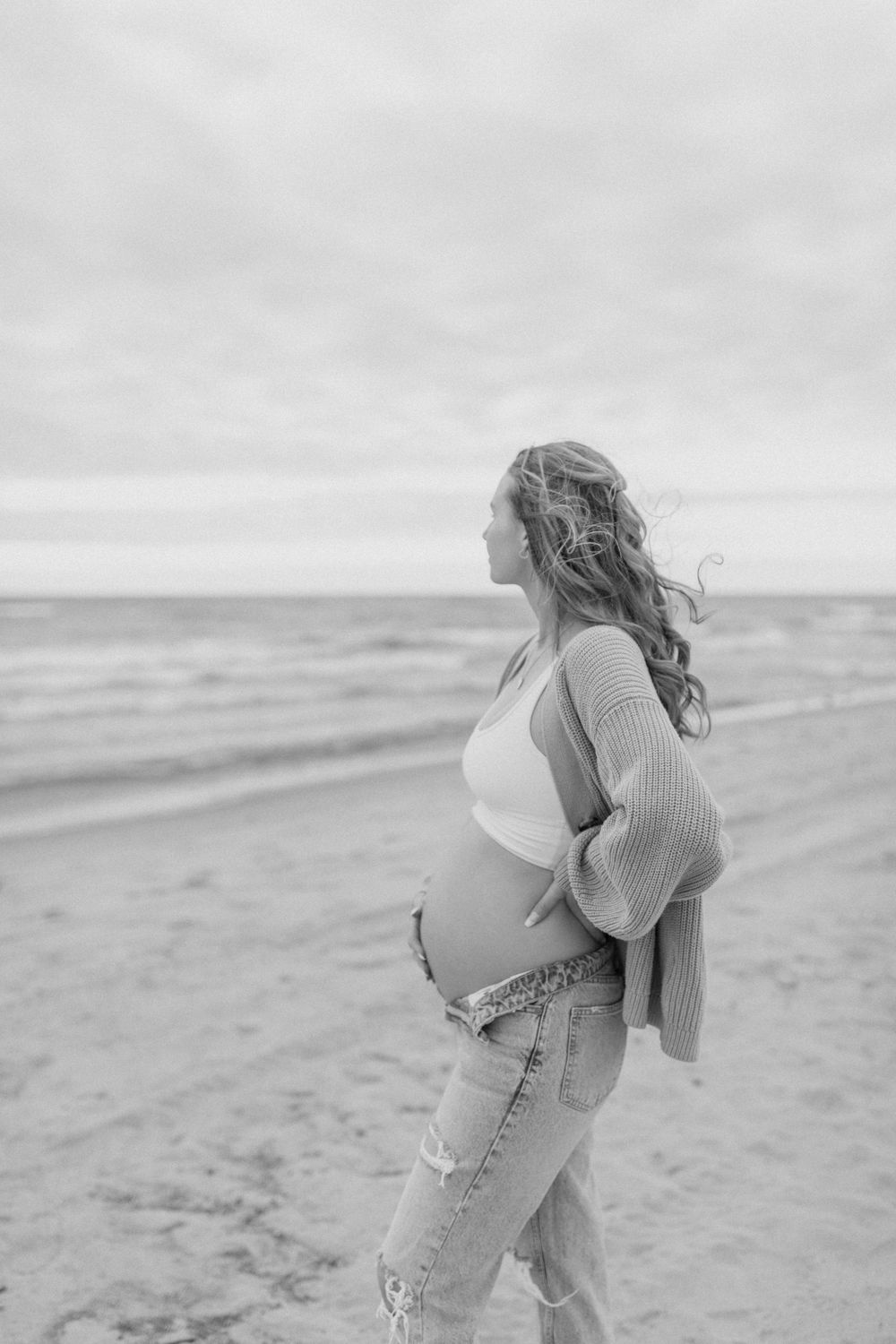 An expectant mother in casual attire stands on a cloudy beach.