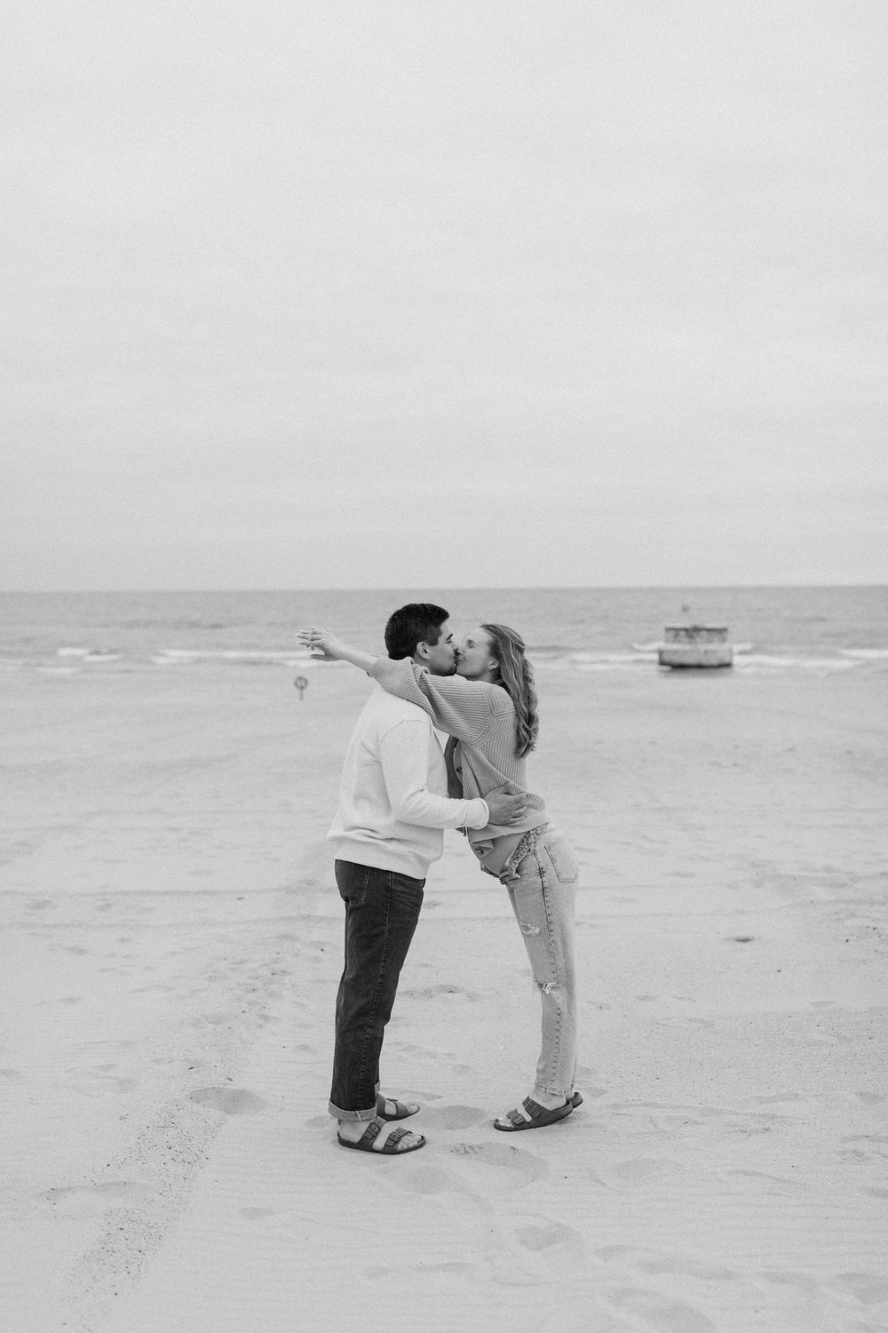 A black and white beach photo of a couple embracing with a boat in the distance.