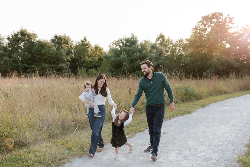 A family walks together on a gravel path through tall grass at sunset.